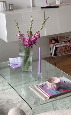 a glass table topped with a vase filled with purple flowers next to a magazine and candle