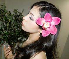 a woman with long black hair holding a pink flower in her right hand and looking down at the ground