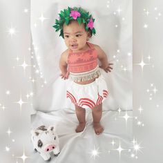 a baby wearing a hula skirt standing next to a stuffed animal on a bed