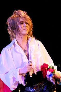 a woman in white shirt and black pants on stage with flowers around her neck, holding a microphone