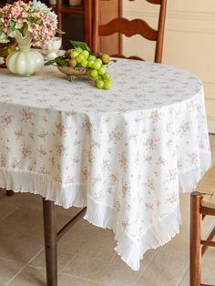 a table with flowers and grapes on it in front of a chair next to a vase