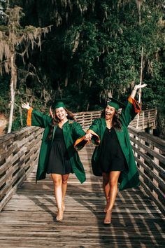 two young women in graduation gowns walking across a wooden bridge with their arms outstretched