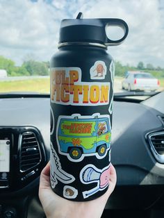 a person holding up a black cup with stickers on it in front of a car