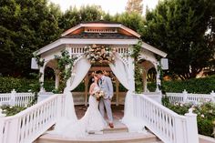 a bride and groom standing in front of a gazebo with flowers on it at their wedding