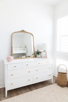 a white dresser topped with a mirror and a baby crib next to a window