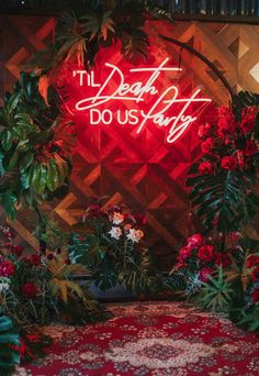 a red neon sign that says the dear do us party surrounded by flowers and greenery