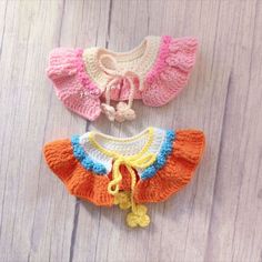 three crocheted baby sweaters are sitting on a wooden surface, one is pink and the other is orange