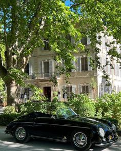 an old black car is parked in front of a large building with trees and bushes