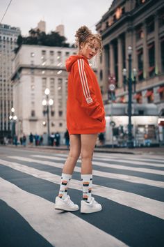 a woman in an orange hoodie is standing on the crosswalk wearing white tennis shoes