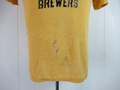 "Vintage 1960s yellow cotton t shirt with black graphics: Richmond Brewers Little League Baseball. Made by Southern Athletic. Size small. Actual measurements are: 38\" around the chest 38\" around the waist 17.5\" shoulder seam to shoulder seam 23\" overall length In \"wrecked\" condition with several holes." Vintage Fitted T-shirt With Letter Print, Fitted Yellow T-shirt With Screen Print, Retro Yellow T-shirt With Screen Print, Vintage Yellow Tops With Screen Print, Vintage Yellow Top With Screen Print, Retro Mustard Cotton Top, Fitted Vintage T-shirt With Screen Print, Vintage Yellow Short Sleeve T-shirt, Retro Yellow Tops With Vintage Print