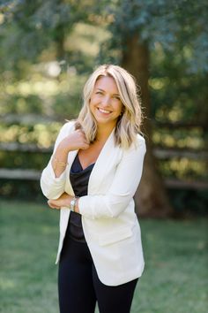 a woman in black pants and a white blazer smiles at the camera with her arms crossed