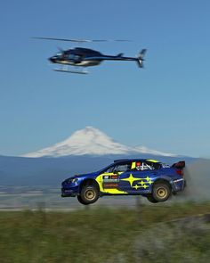 a helicopter flying over a blue car in the air with mountains in the back ground