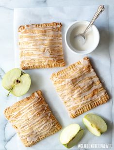 apple pies with icing and sliced apples on a white marble countertop next to a cup of yogurt