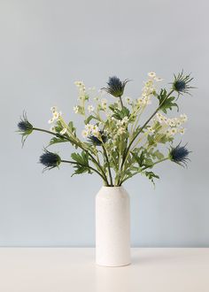 a white vase filled with lots of flowers on top of a table next to a gray wall