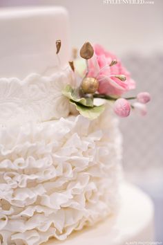 a white wedding cake with ruffled layers and pink flowers on the top, sitting on a table