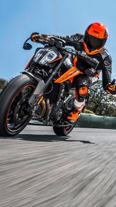 a man riding on the back of an orange and black motorcycle down a street with trees in the background