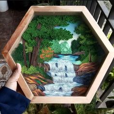 a person holding up a painting in front of a wooden frame with a waterfall and trees on it