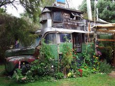 an old bus is covered in flowers and plants next to a wooden structure with a house on top