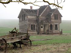 an old wooden house sitting in the middle of a field next to a horse drawn wagon