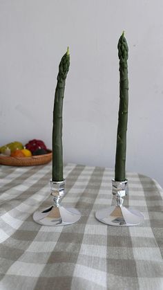 two green candles sitting on top of a checkered table cloth with fruit in the background