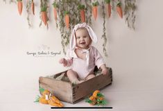 a baby sitting in a wooden crate with carrots hanging from the wall behind it