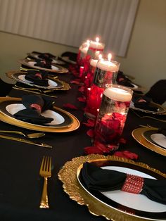 the table is set with black and gold plates, silverware, red roses and candles