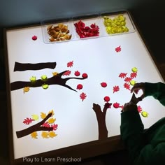 a child is playing with fall leaves on a white lit table in front of trees