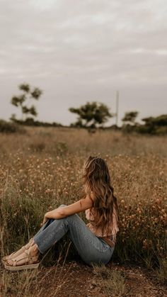 a woman sitting in the middle of a field with her hands on her knees and looking off into the distance