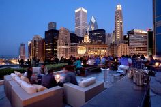 a group of people sitting at tables on top of a roof with city lights in the background