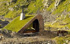 a small house with a green roof on the side of a mountain covered in grass