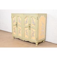 an antique cabinet painted in yellow and white with floral designs on the doors, stands against a brick wall