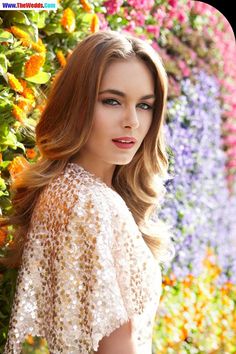 a beautiful woman with long hair standing in front of colorful flowers wearing a white dress