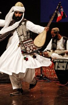 two men in traditional garb playing musical instruments on stage with other musicians behind them