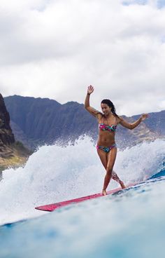 a woman riding a surfboard on top of a wave in the ocean with mountains behind her