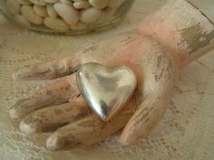 a hand holding a silver heart on top of a white table cloth next to a jar filled with beans