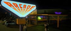 people standing in front of an arcade sign at night with cars parked outside the building