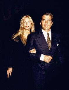 a man in a suit and tie standing next to a blonde haired woman with her arms crossed