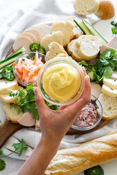 a person holding a jar filled with mayonnaise next to bread and veggies