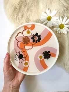 a person holding a plate with flowers painted on it next to a stuffed animal and some daisies