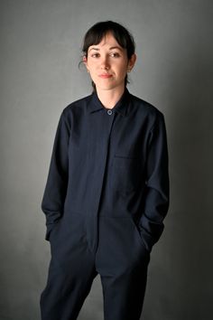 a woman standing with her hands in her pockets and looking at the camera while wearing a black jumpsuit