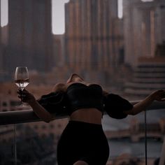 a woman holding a glass of wine on top of a balcony next to a city