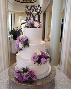 a three tiered white cake with purple flowers on the top and bottom, sitting on a table in front of a chandelier