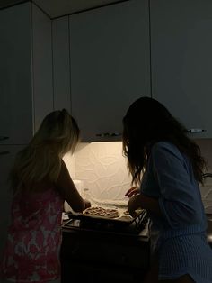 two girls are cooking in the kitchen at night, one is pulling something out of the oven