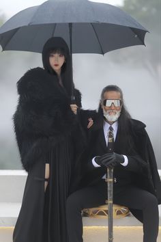 two people sitting on a bench with an umbrella in front of them, both wearing black clothing