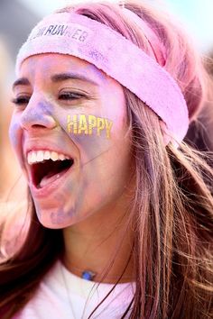 a girl with her face painted in the colors of yellow and pink is laughing while wearing a headband that says happy