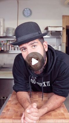 a man with a beard and top hat leaning on a table in a kitchen looking at the camera