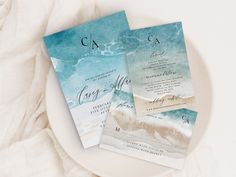 two wedding cards sitting on top of a white plate next to each other with watercolor designs