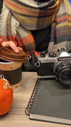 a person holding a coffee cup next to a camera and a pumpkin on a table