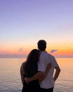 a man and woman standing next to each other near the ocean at sunset or sunrise