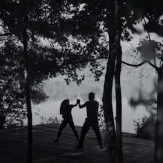 two people are dancing on a wooden platform in the woods near water, with trees around them